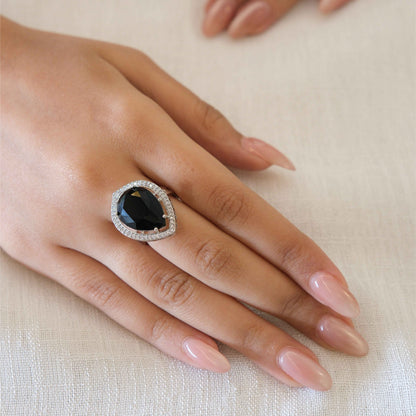 A close-up of a hand wearing a teardrop-shaped ring with a dark oval center stone and a silver-toned halo, set against a soft beige fabric background.