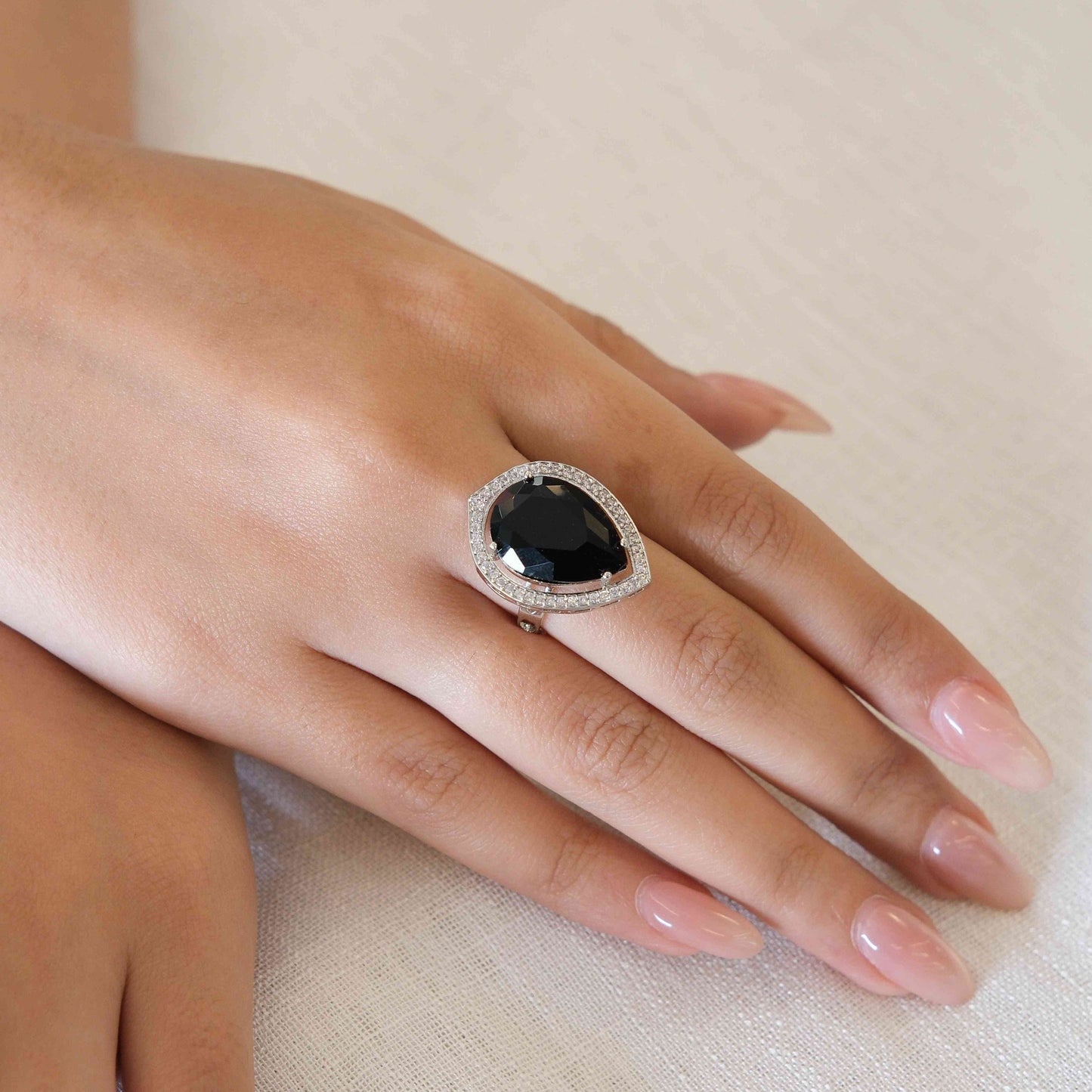 A close-up of a hand wearing a silver-toned teardrop ring with a large black oval center stone and a halo of small clear stones, resting on a light beige fabric surface.
