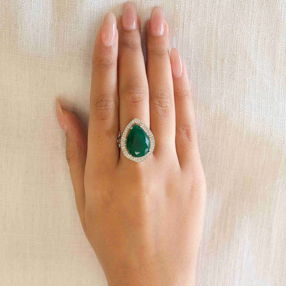 A hand with manicured nails wearing a teardrop-shaped green gemstone ring set in silver, resting on a textured beige fabric background.