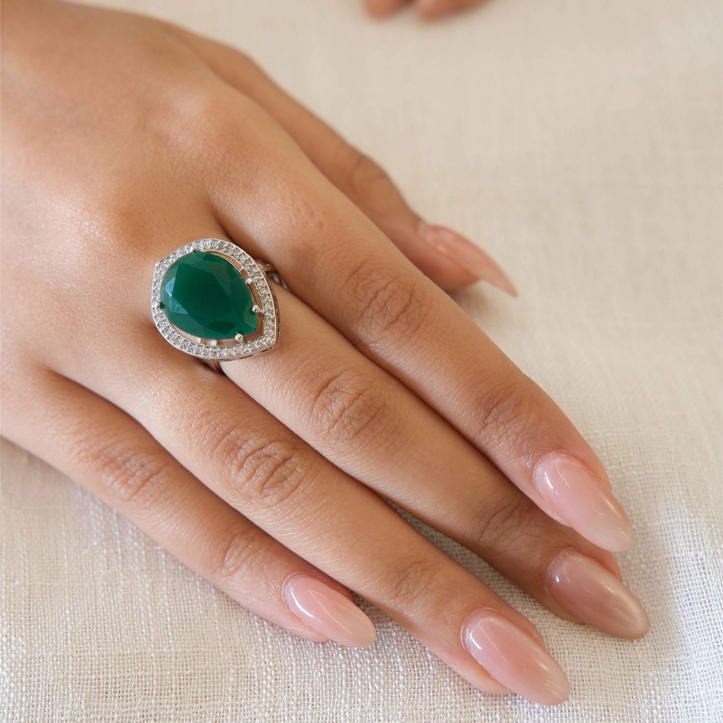 A close-up of a hand wearing a ring with a large green teardrop-shaped center stone surrounded by a halo of small clear stones, set against a soft beige fabric background.