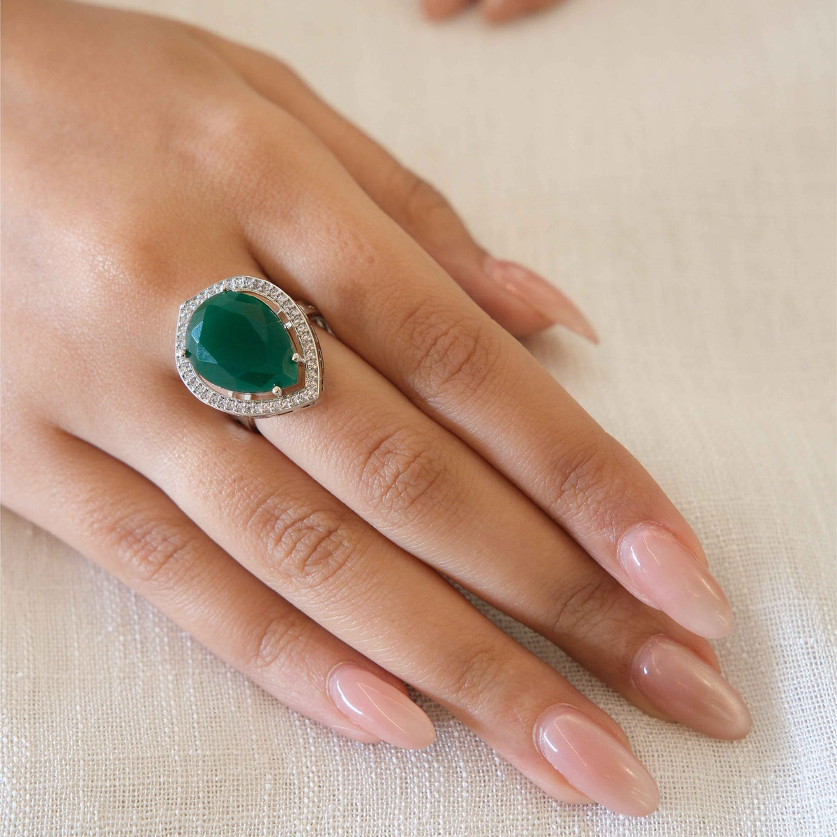 A close-up of a hand wearing a ring with a large green teardrop-shaped center stone surrounded by a halo of small clear stones, set against a soft beige fabric background.
