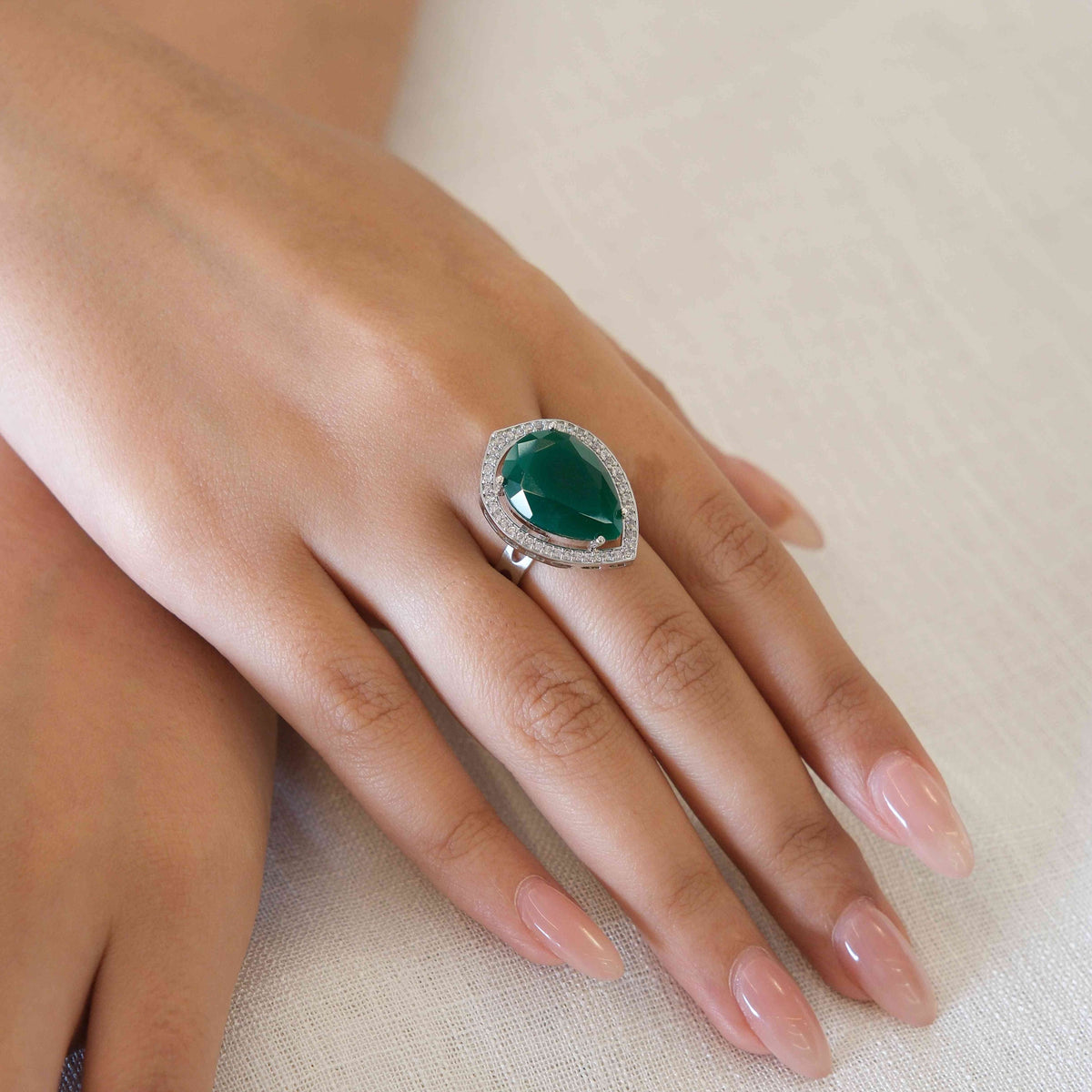 A close-up of a hand wearing a large teardrop-shaped green gemstone ring with a silver halo and small clear stones, resting on a light beige fabric surface.