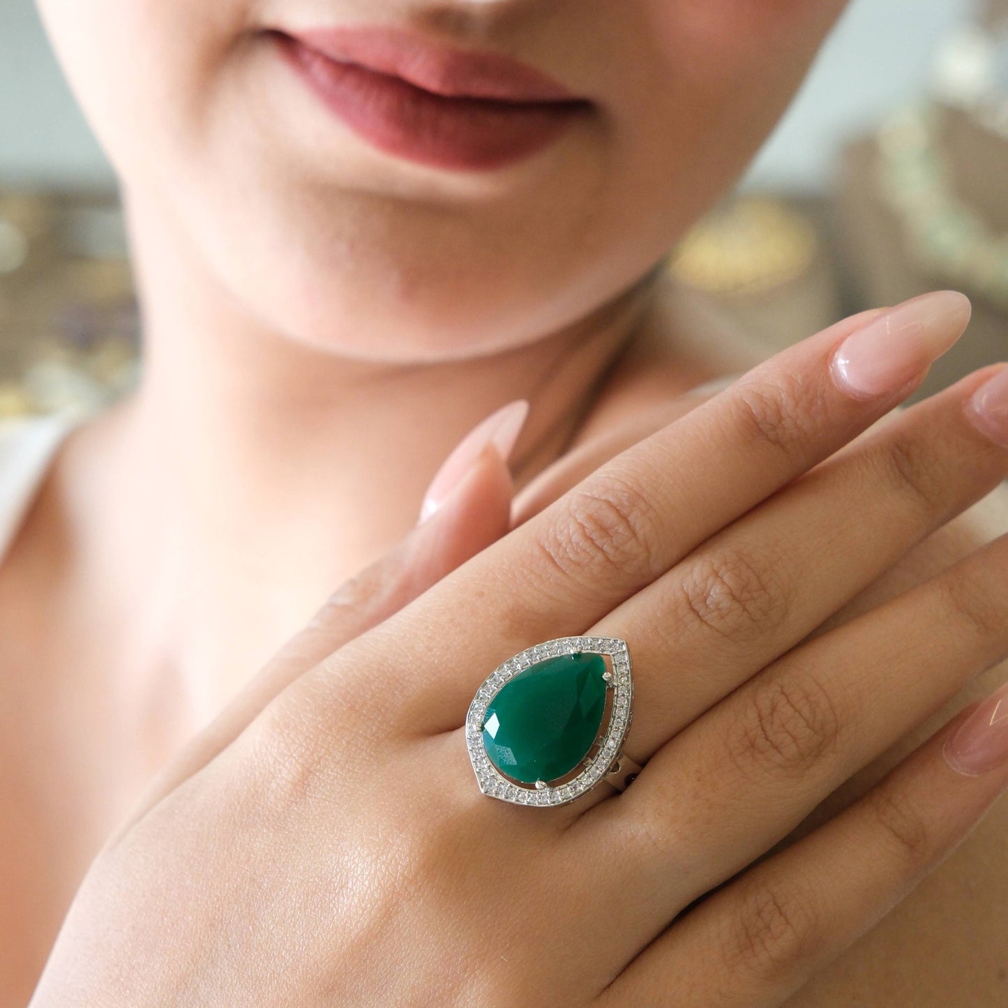 A close-up lifestyle shot of a woman wearing a teardrop-shaped green gemstone ring with a halo of small clear stones on her finger.
