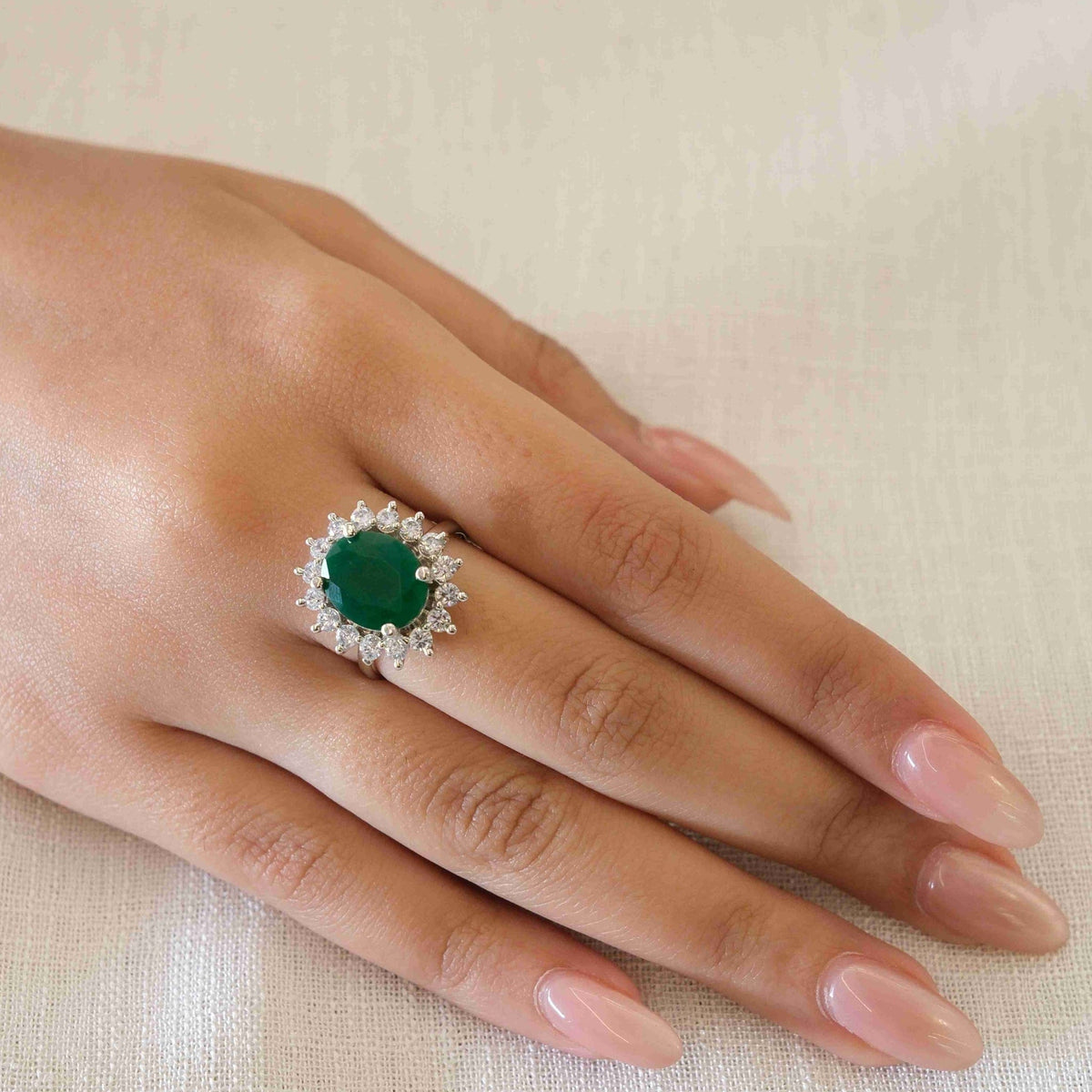 A close-up of a hand wearing a silver ring with a round green emerald-cut center stone surrounded by small clear stones, resting on a textured off-white fabric background.
