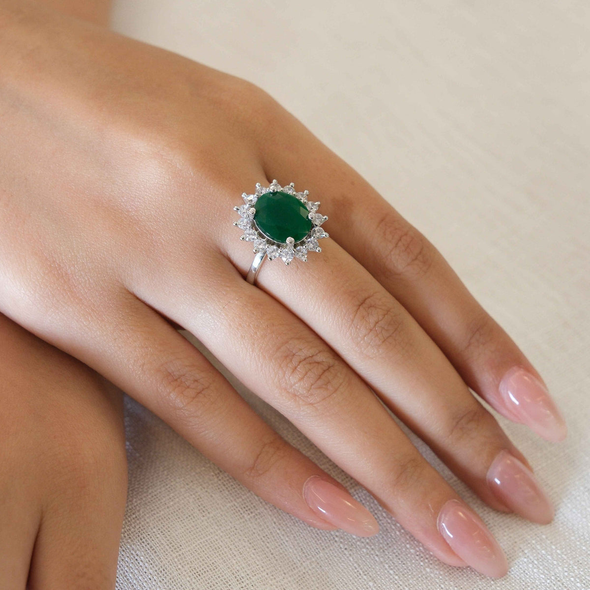 A silver ring featuring a round green emerald-cut center stone surrounded by a halo of small clear stones, shown worn on a hand resting on a light beige textured fabric.