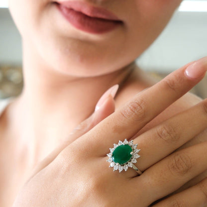 A close-up lifestyle shot of a woman's hand wearing a silver ring with a round green emerald-cut center stone surrounded by a halo of small clear stones.