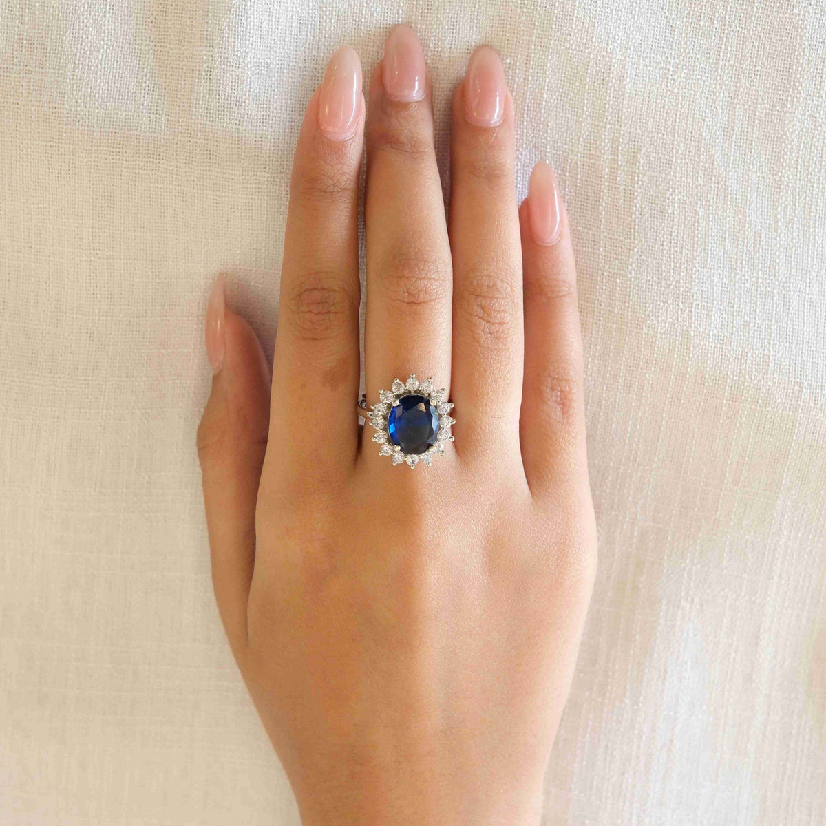 A silver ring with a central oval-cut dark blue gemstone surrounded by small clear stones, worn on a person's hand against a light beige textured fabric background.