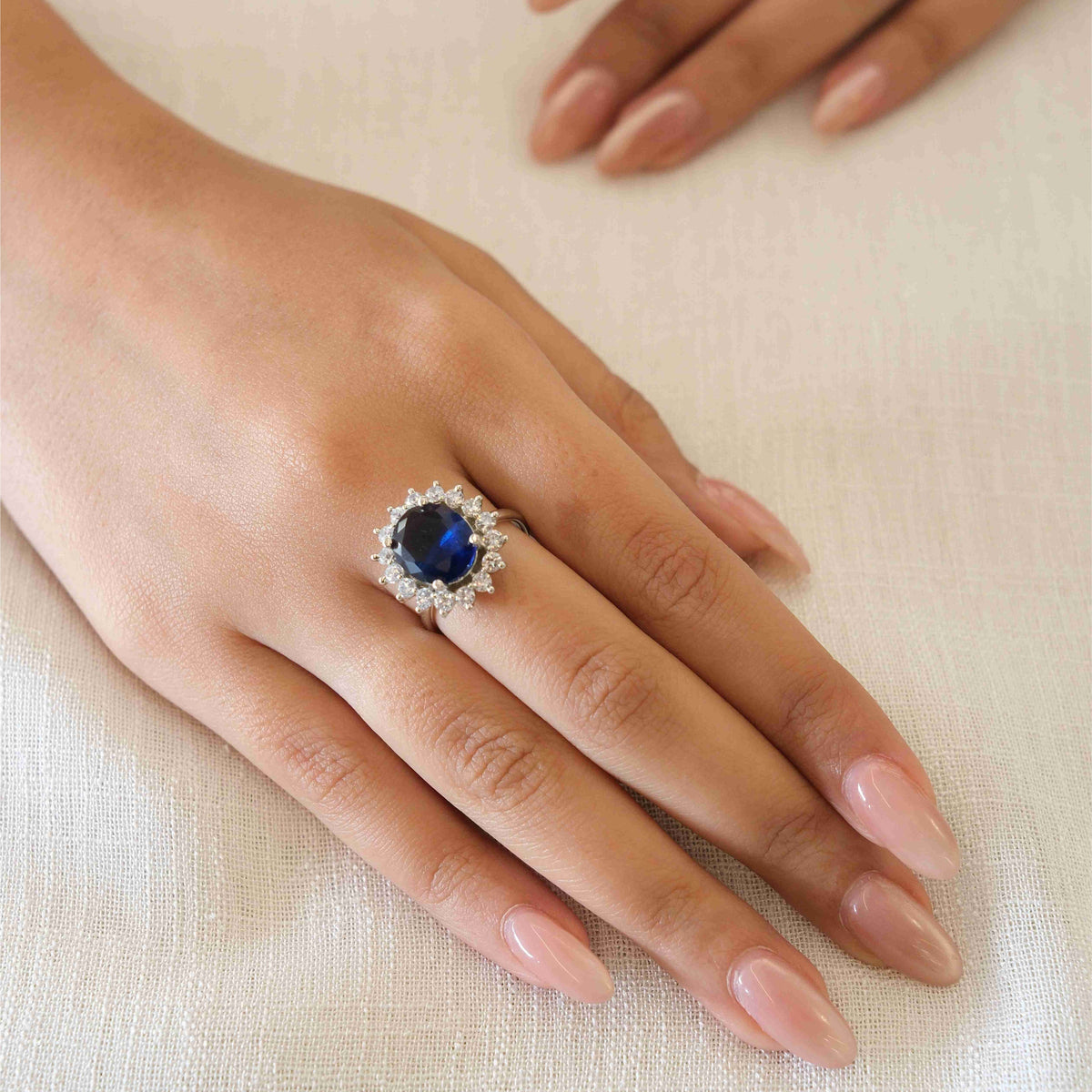 A silver-toned ring featuring a round deep blue gemstone surrounded by small clear stones, worn on a hand resting on a textured cream fabric background.