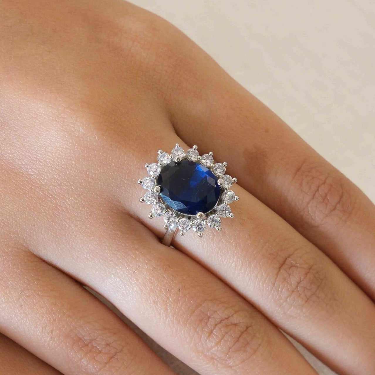 A close-up of a hand wearing a silver ring with a large oval blue gemstone surrounded by a halo of small clear stones.