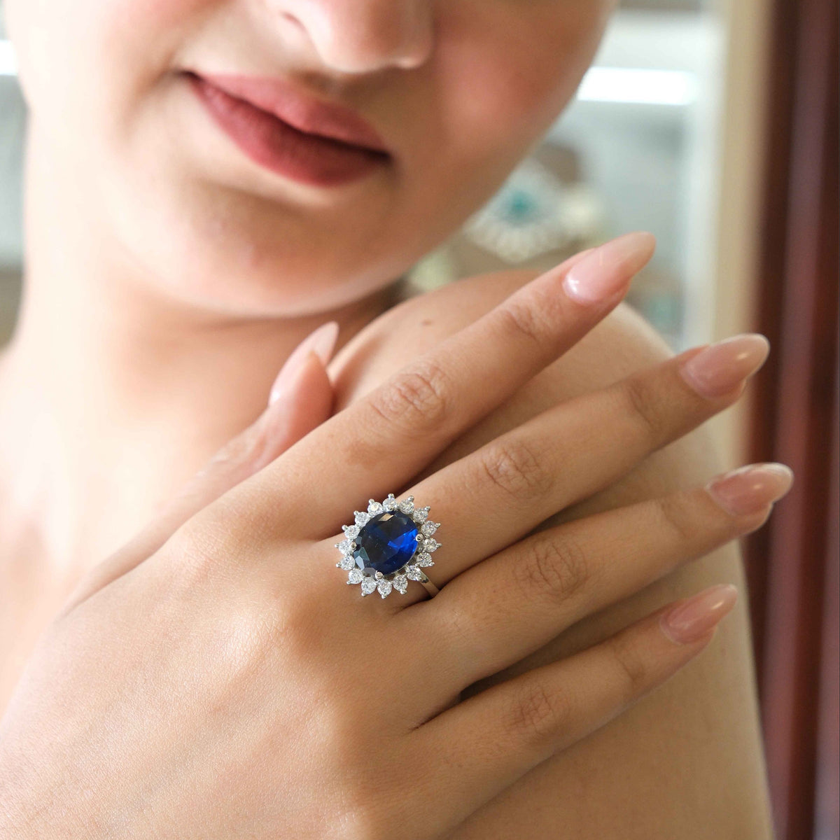 A close-up lifestyle shot of a woman wearing a silver ring with a large oval-cut blue sapphire and a halo of small clear stones.