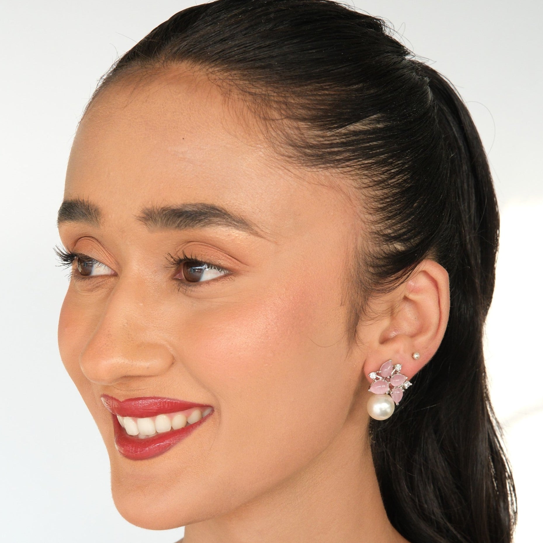 A woman with dark hair pulled back wears a small cluster earring with pink stones and a pearl, shown in a close-up portrait against a light background.
