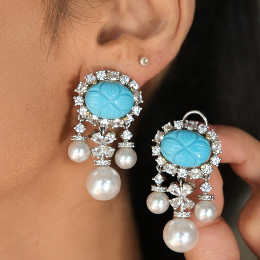A close-up of a person wearing and holding a pair of dangling earrings. The earrings feature oval light blue stone centers surrounded by clear rhinestones, with white pearl drops below.