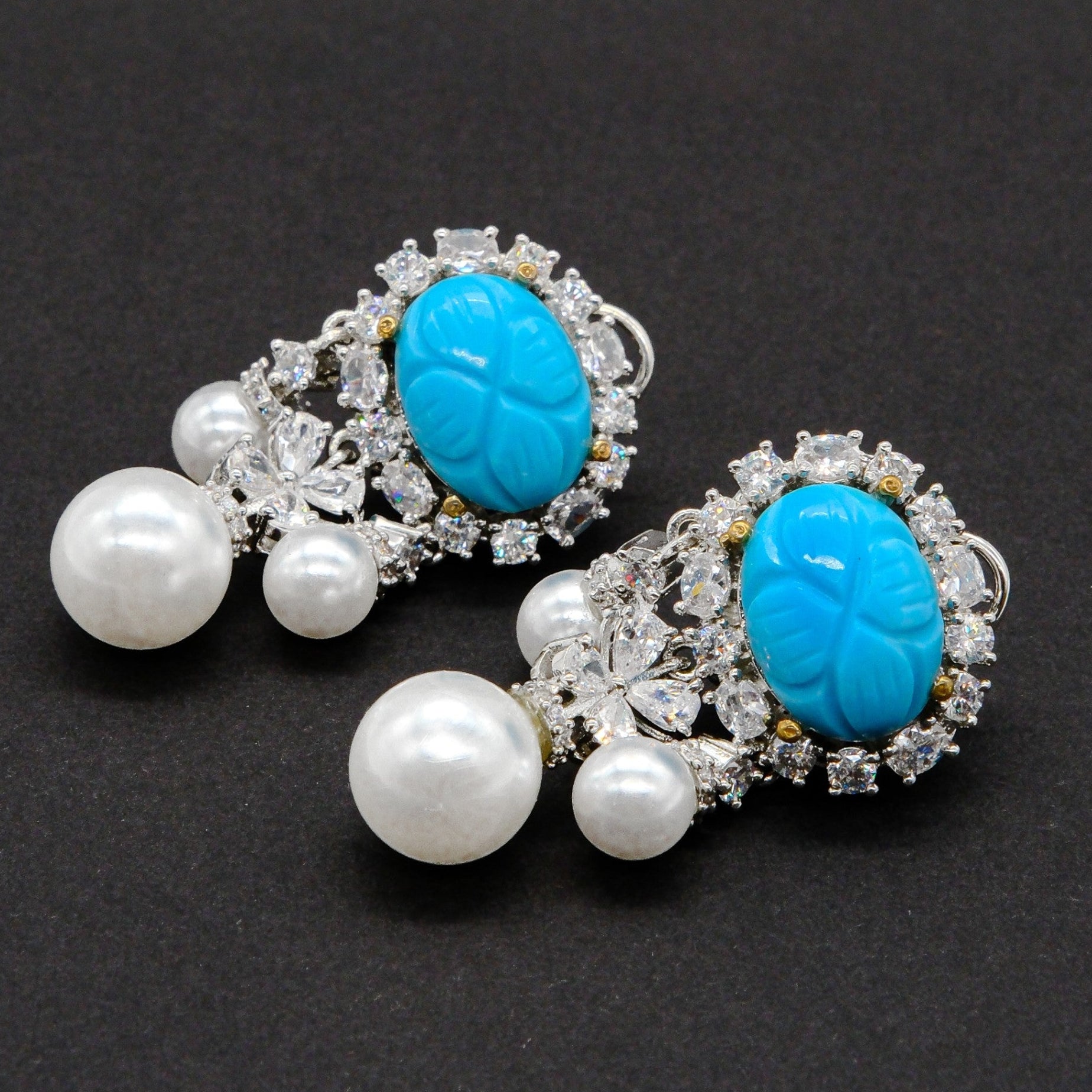 A pair of silver-toned statement earrings featuring a carved turquoise cabochon surrounded by clear crystals and dangling white faux pearls, set against a dark textured background.